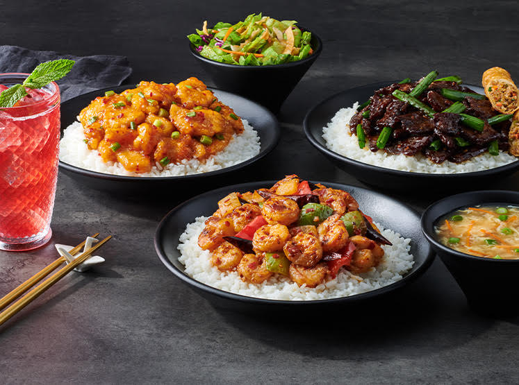 three bowls of sautéed chicken, shrimp, and beef over white rice. A bowl of salad and soup. Large glass of juice with mint sprig. Chopsticks on holder